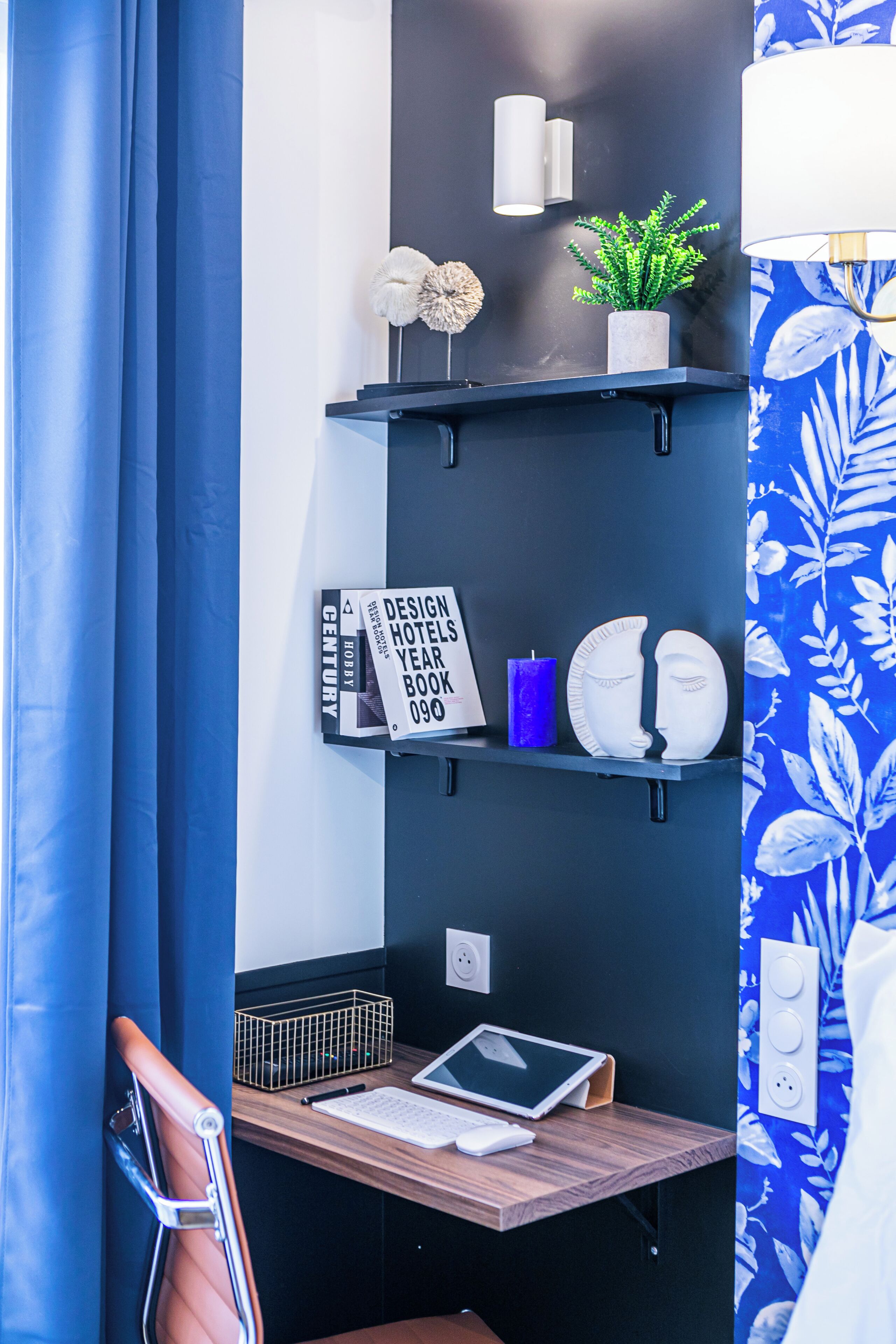 Une chambre d’hôtel moderne avec un mur d’accent bleu foncé, un bureau en bois avec un ordinateur portable, une chaise et une étagère ornée d’objets décoratifs.