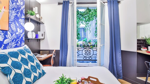 A modern hotel room with a bed, breakfast tray, and a view of a balcony with blue curtains. - WS Louvre - Saint-Germain (Paris)