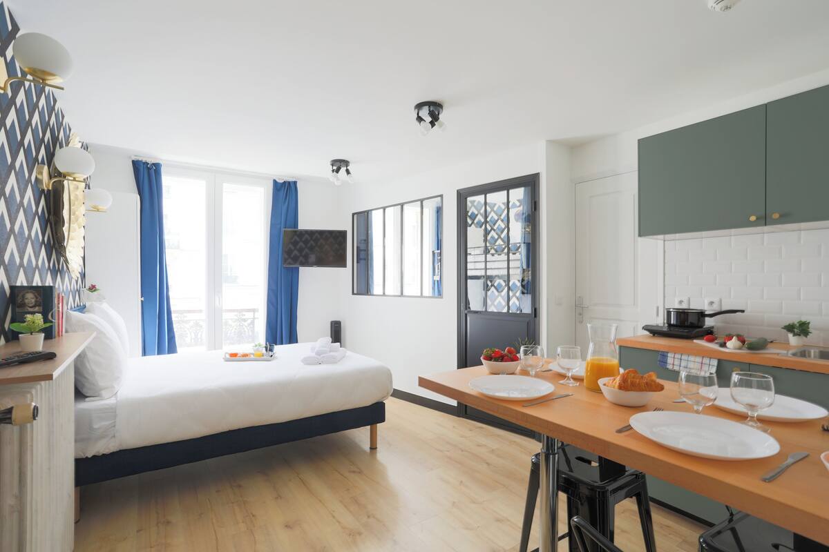 A modern hotel room with a bed, a dining table set for breakfast, a kitchen area, and a balcony with blue curtains.
