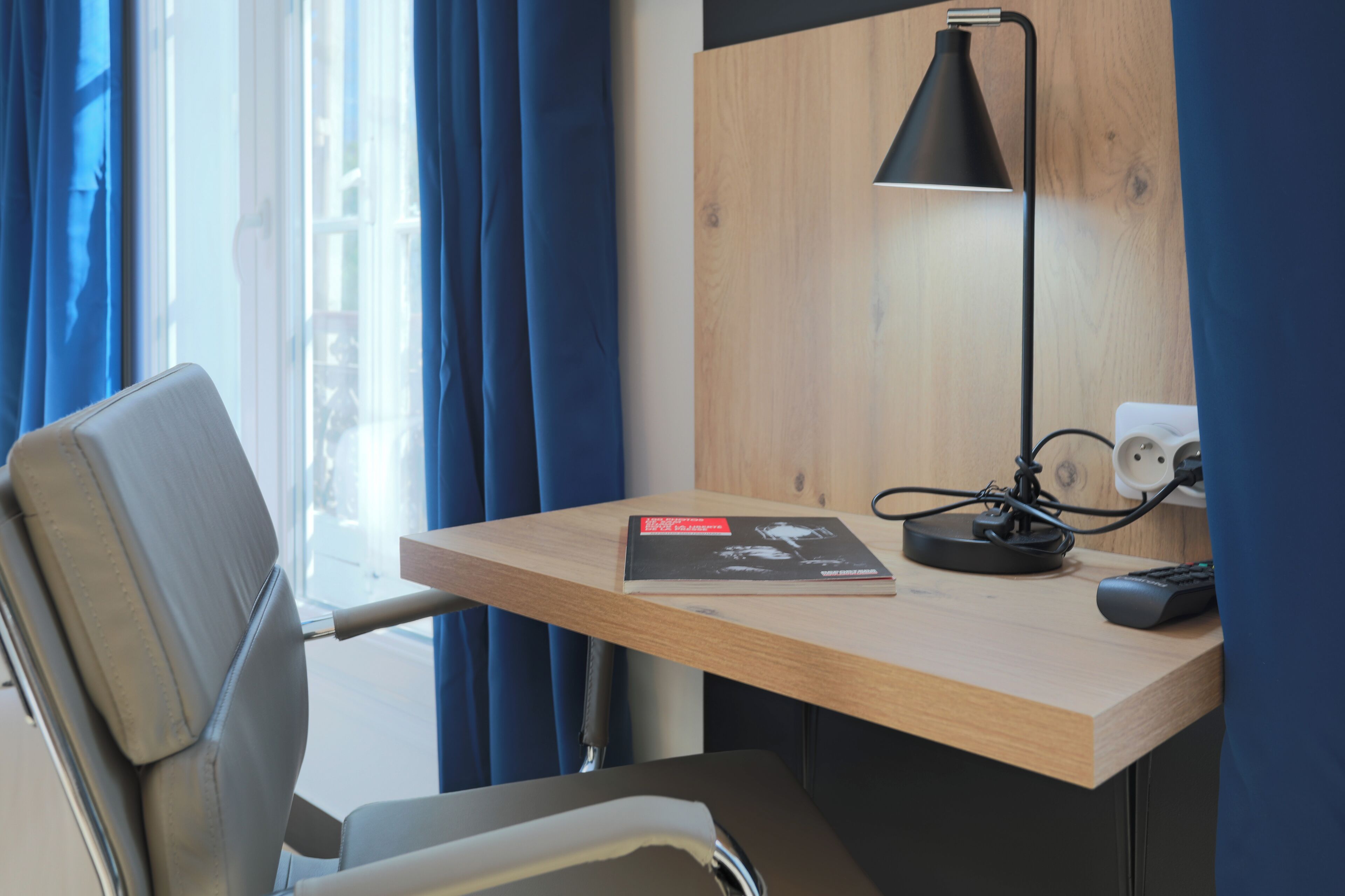 A hotel room with a wooden desk, a black desk lamp, a remote control, and a book.