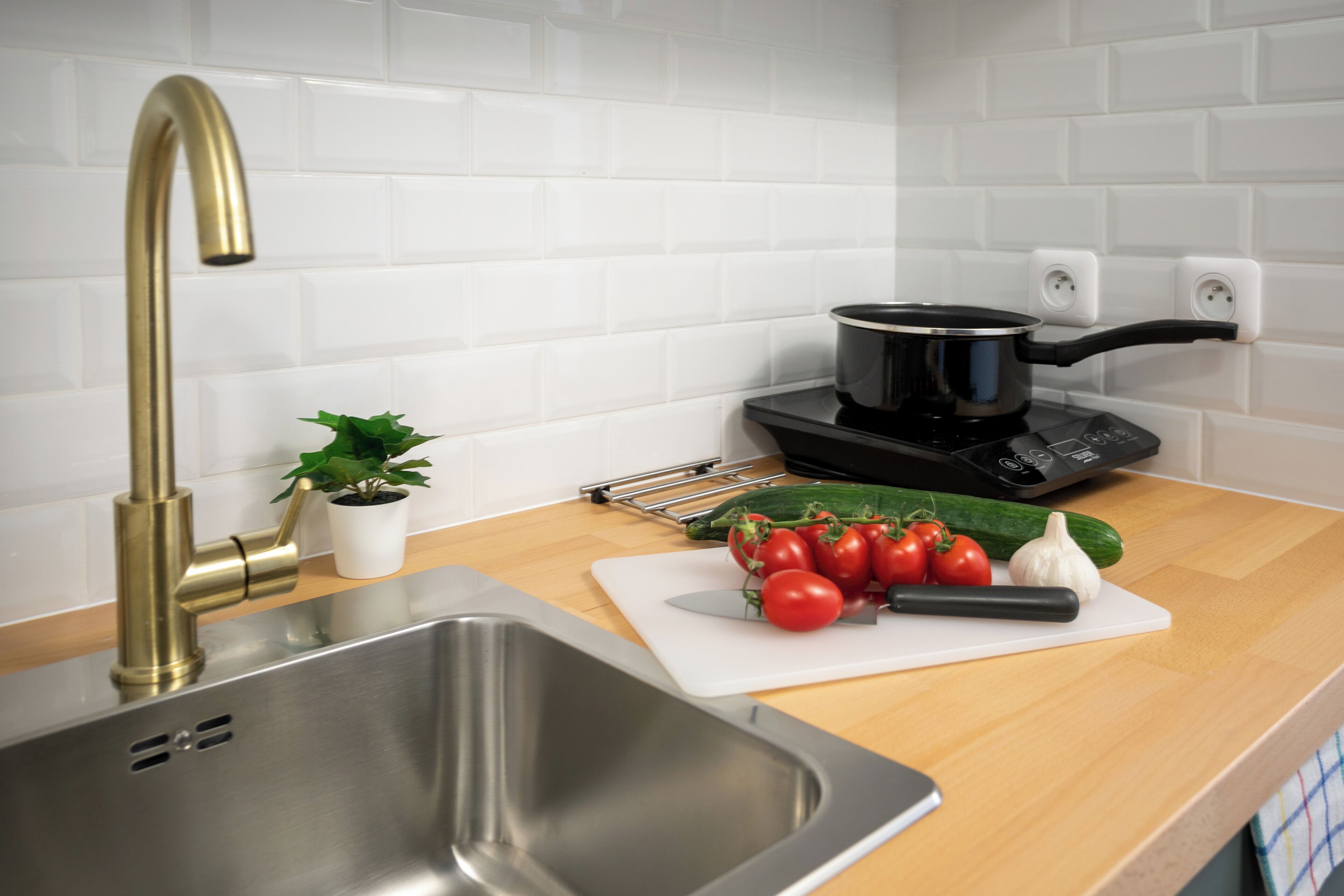 A kitchen with a stainless steel sink, a gold faucet, a black induction cooktop, and fresh vegetables on the counter.