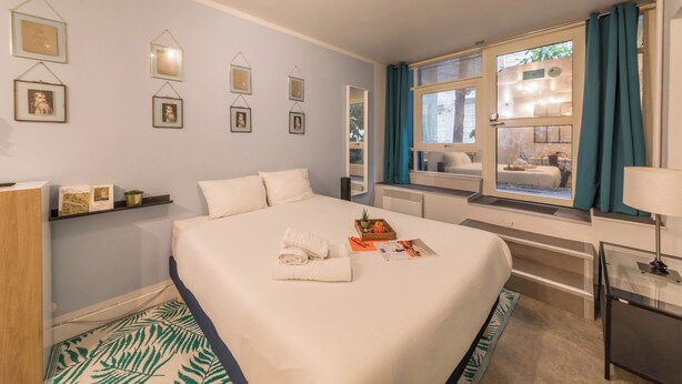 A hotel room with a large bed, a window with curtains, a bedside table with a lamp, and wall-mounted pictures. - WS Montmartre - Sacré Coeur (Paris)