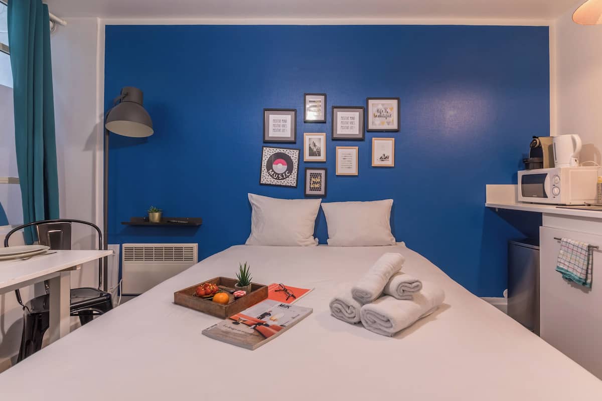 A hotel room with a blue accent wall, a bed with white bedding, a bedside table with a tray of fruits and a magazine, a microwave, and a small shelf with a plant.