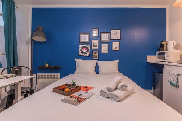 A hotel room with a blue accent wall, a bed with white bedding, a bedside table with a tray of fruits and a magazine, a microwave, and a small shelf with a plant. - (Paris)