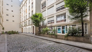 A cobblestone street flanked by multi-story buildings with balconies, a tree-lined sidewalk, and a building entrance with a blue door. - WS Montmartre - Sacré Coeur (Paris)