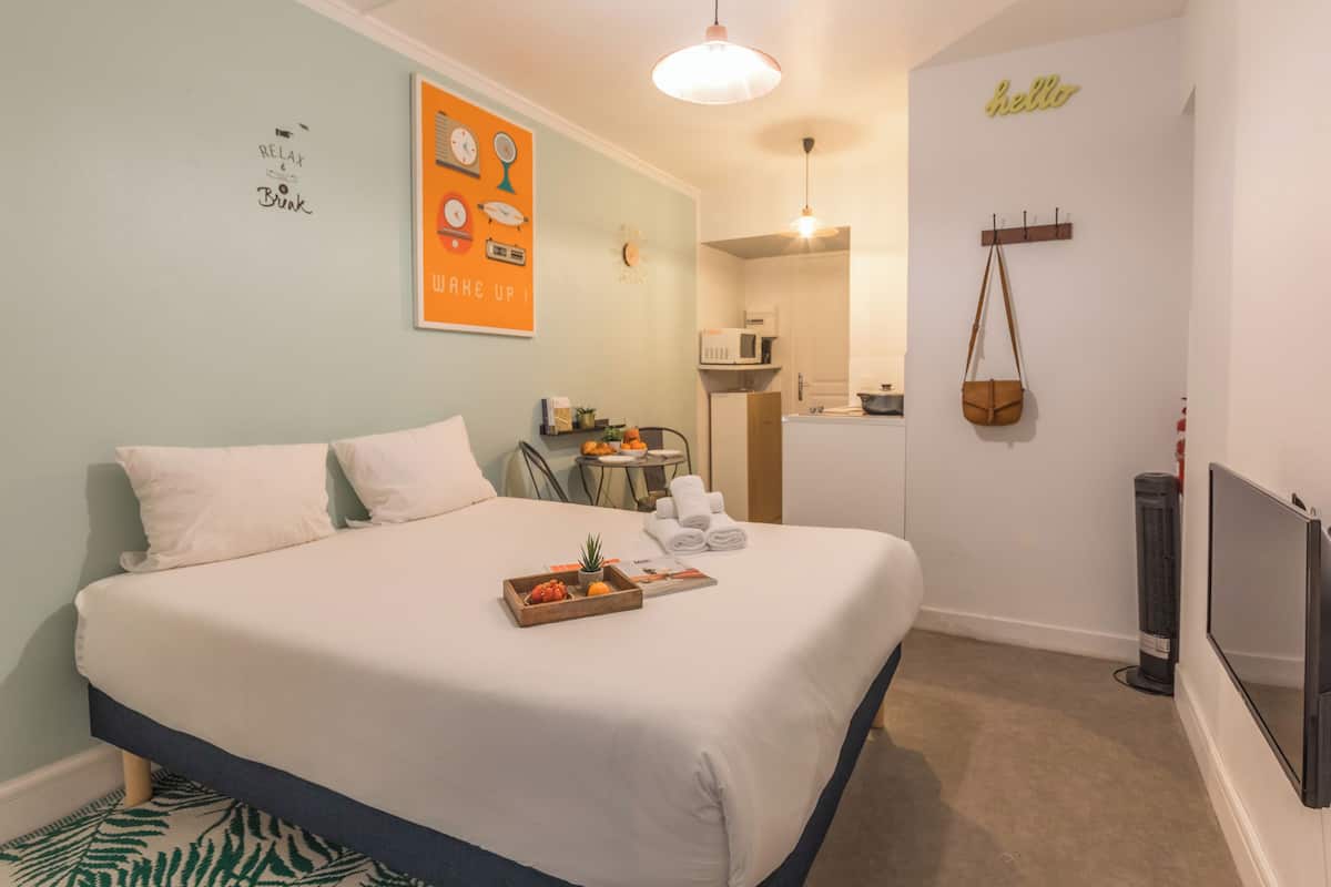 A neatly made bed with a tray of fruits and a small plant, a microwave, and a hanging bag on the wall.