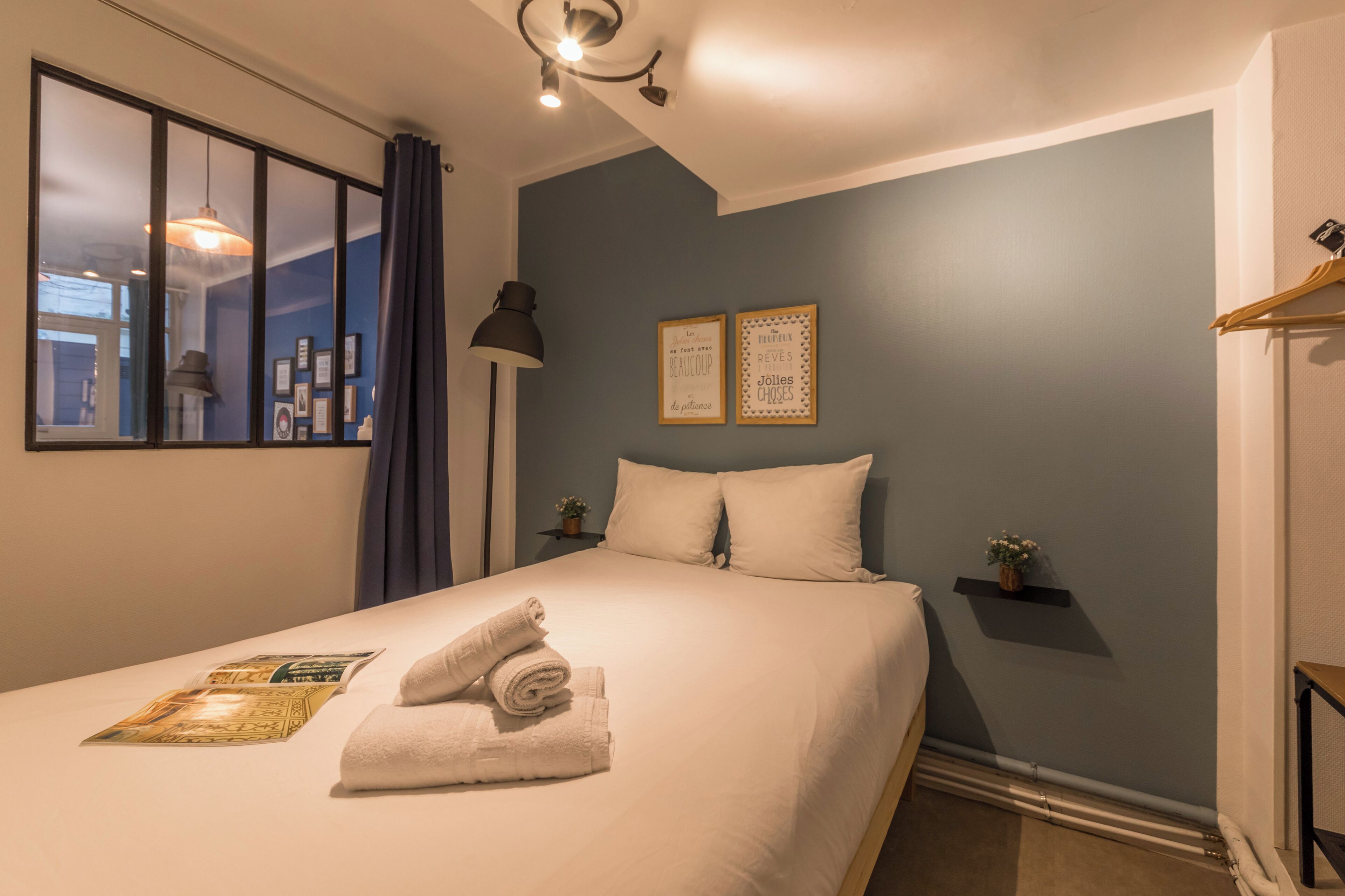 A neatly made bed with rolled towels and a magazine, a bedside lamp, and two framed quotes on the wall.