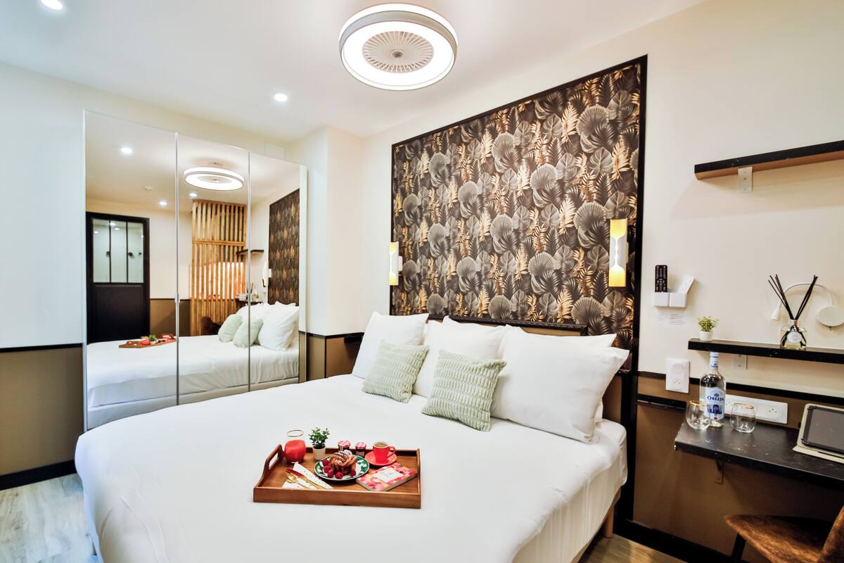 A modern hotel room with a large bed, a bedside table with a tray of breakfast, a large mirror, and a decorative wall panel.