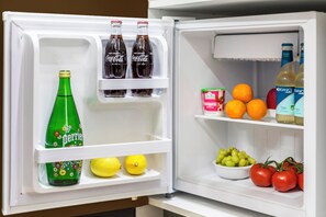Un réfrigérateur ouvert, dont les étagères contiennent des bouteilles de Coca-Cola, de Perrier et d’eau tonique, ainsi que des oranges, des citrons, des raisins et des tomates.