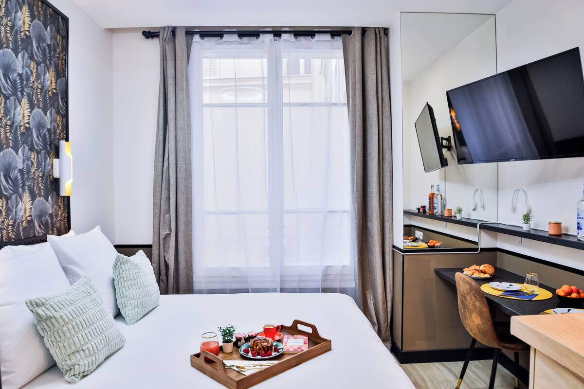 A hotel room with a bed, breakfast tray, a desk, a TV, and a window with curtains.