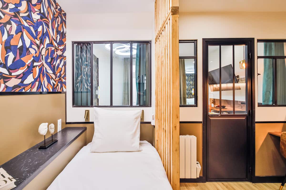 A modern hotel room with a bed, a wooden partition, a desk with a lamp, and a colorful abstract wall.