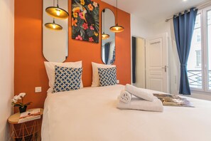 Cama bem arrumada com lençóis brancos e toalha enrolada, duas almofadas estampadas, um espelho fixado na parede e uma pintura floral tropical.