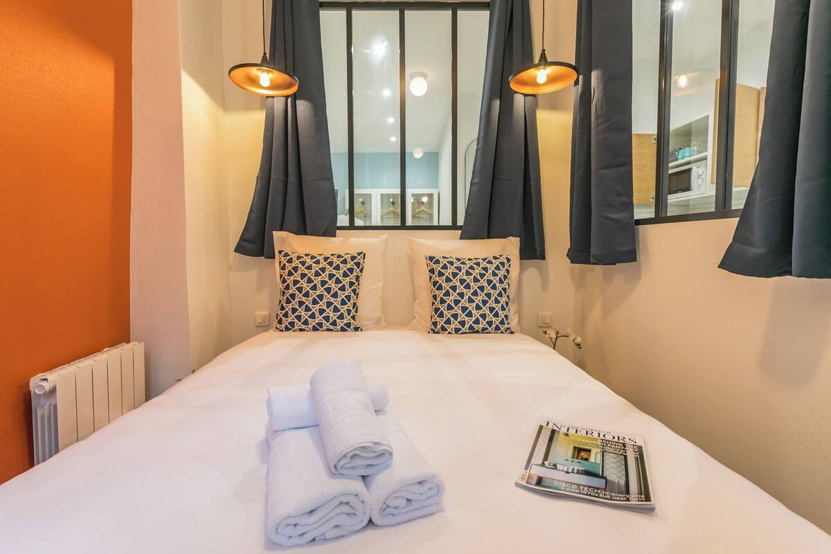 Una habitación de hotel con una cama bien hecha, dos almohadas estampadas, una revista sobre la cama y una ventana con cortinas.
