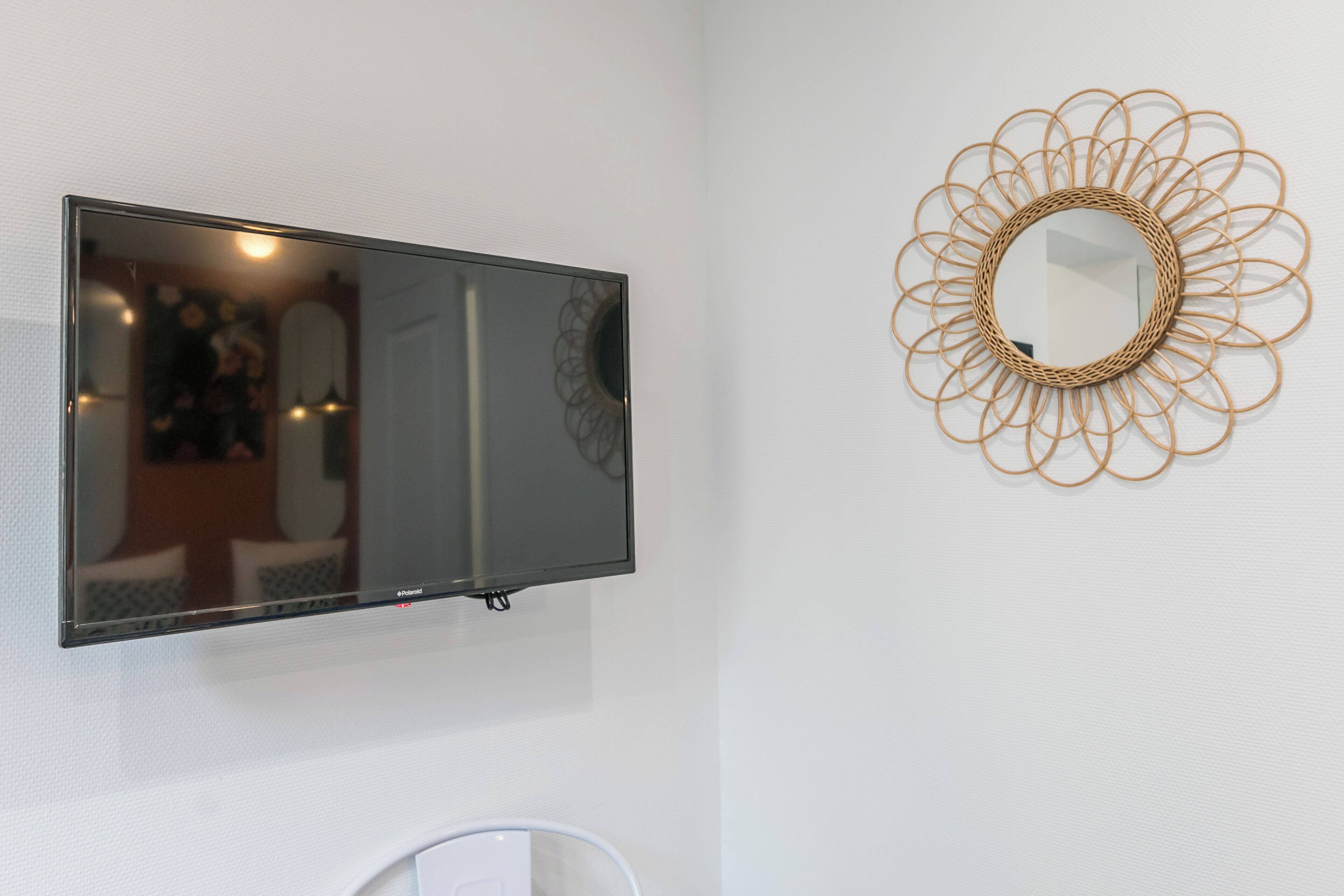 Uma televisão de tela plana fixada em uma parede branca, um espelho decorativo com moldura circular e uma cadeira branca.
