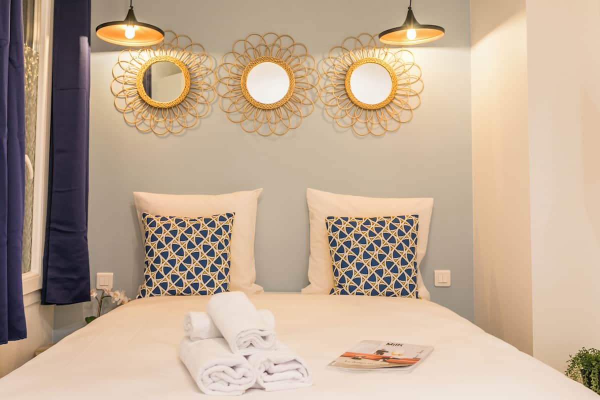 Una cama bien hecha con toallas enrolladas y una revista, tres espejos fijados a la pared y dos almohadones estampados.
