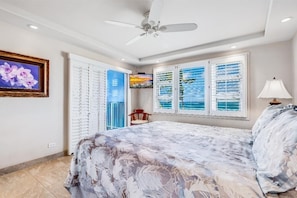 A bedroom with a bed, a ceiling fan, a window with blinds, a framed picture on the wall, and a lamp.