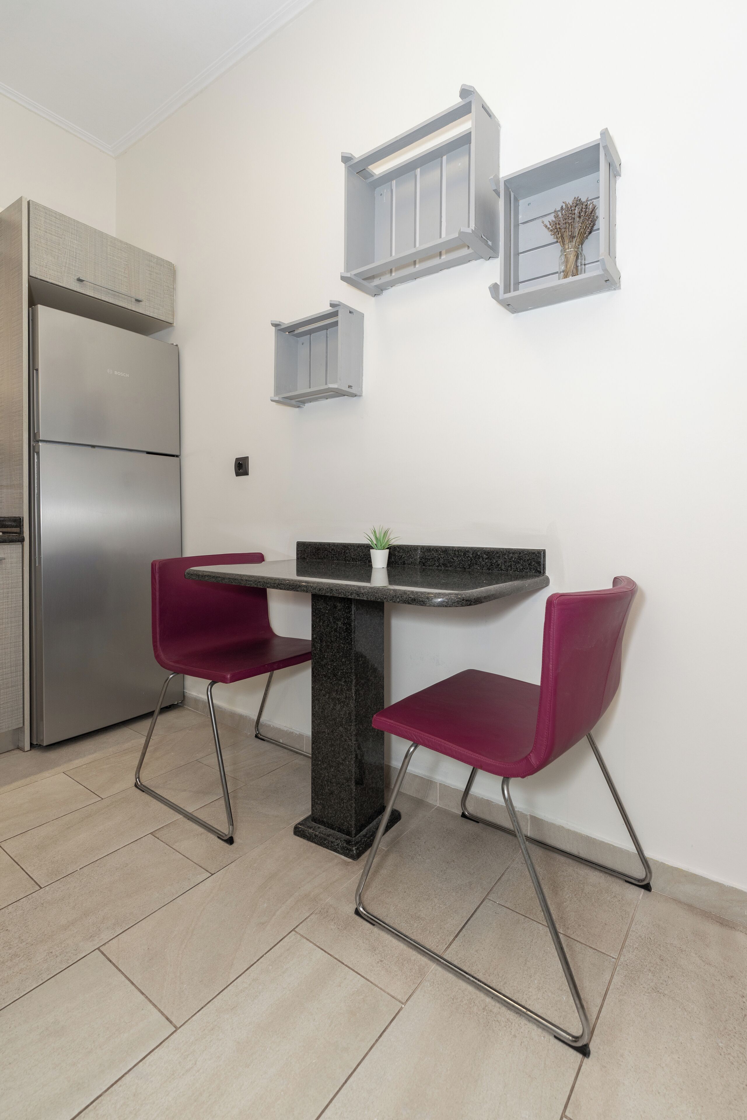 A compact kitchen with a stainless steel refrigerator, a granite countertop, and two red chairs.