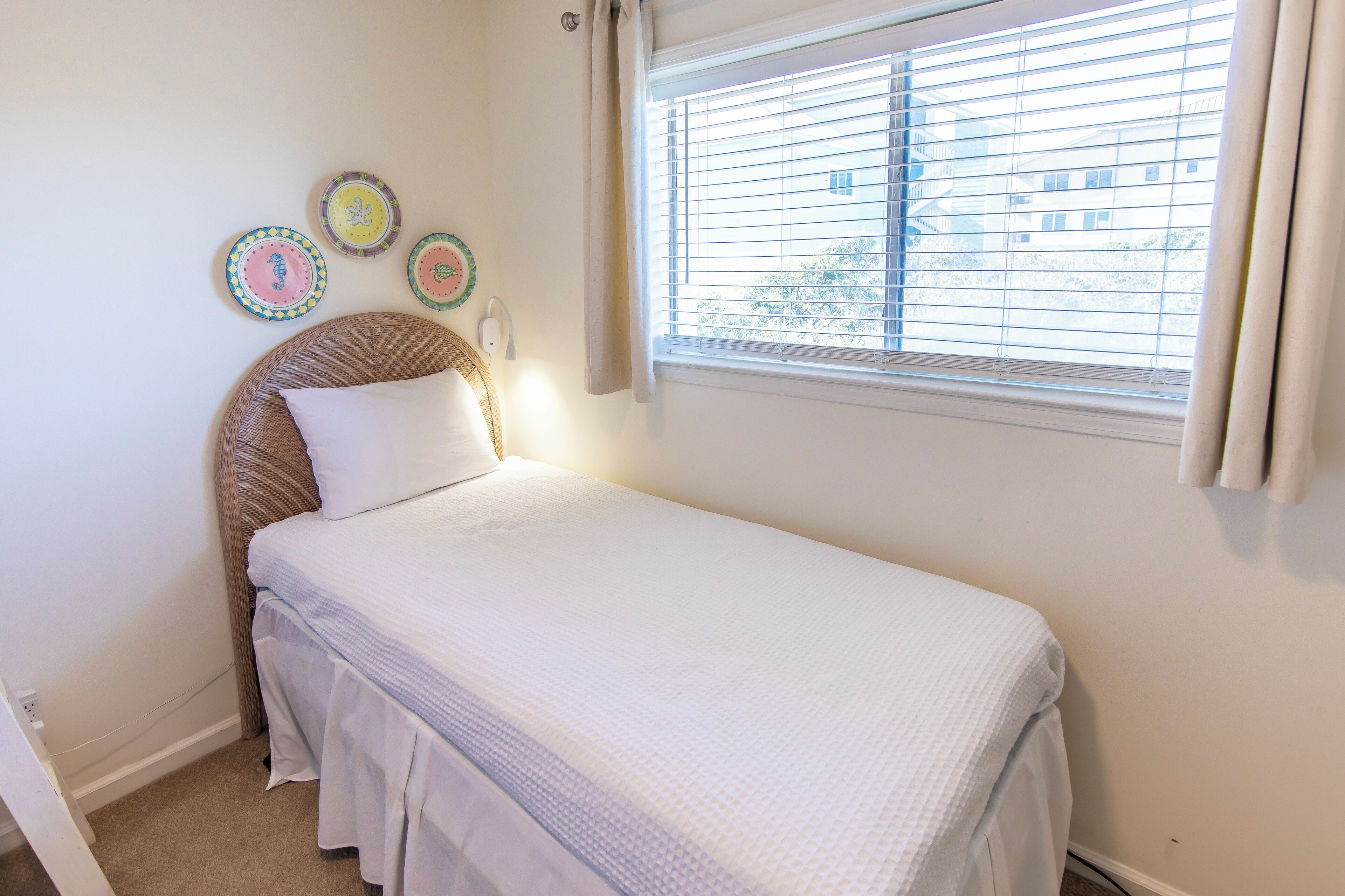 A small, neatly made bed with a white comforter and a wicker headboard. There are three decorative plates on the wall above the bed.