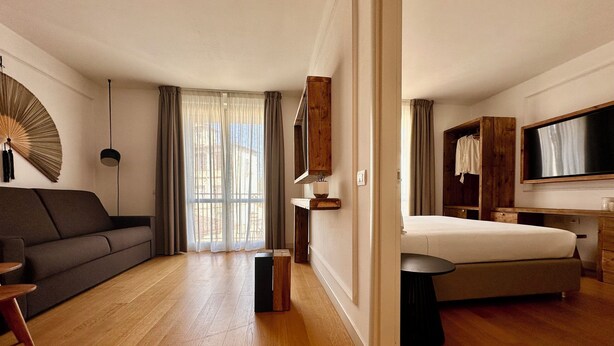 Habitación de hotel moderna con piso de madera, sofá, cama, televisor y una ventana grande con cortinas.