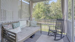 A covered porch with a cushioned seating area and a rocking chair, overlooking a green landscape. - Spacious 4-bed 3.5-bath Townhome in Long Neck with AC, Swimming Pool & Game Room (Long Neck)