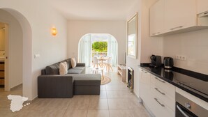 Cozinha moderna com armários brancos e bancada preta, área de jantar com mesa e cadeiras redondas, e sala de estar com sofá cinza e lareira.