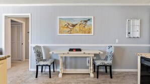 A dining area with a table and two chairs, a painting on the wall, and a window with blinds.
