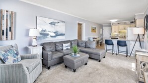 A modern living room with a grey sectional sofa, a small ottoman, and a coffee table. There is a kitchen area with a bar stool and a large painting on the wall.