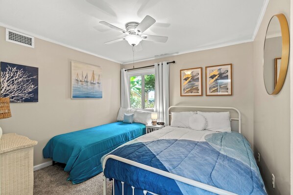 A bedroom with two beds, a ceiling fan, a window with curtains, and artwork on the walls.