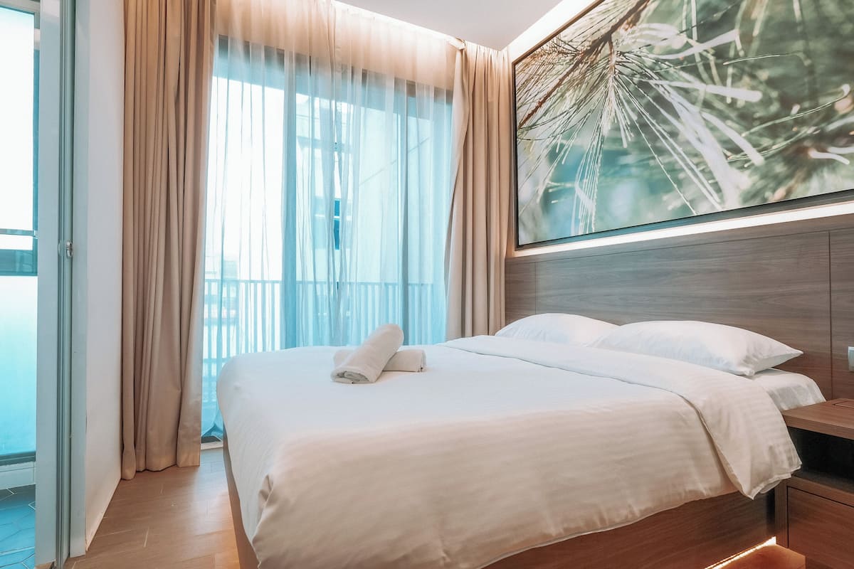 Quarto de hotel moderno com cama grande, cabeceira de madeira e uma janela ampla com cortinas leves.