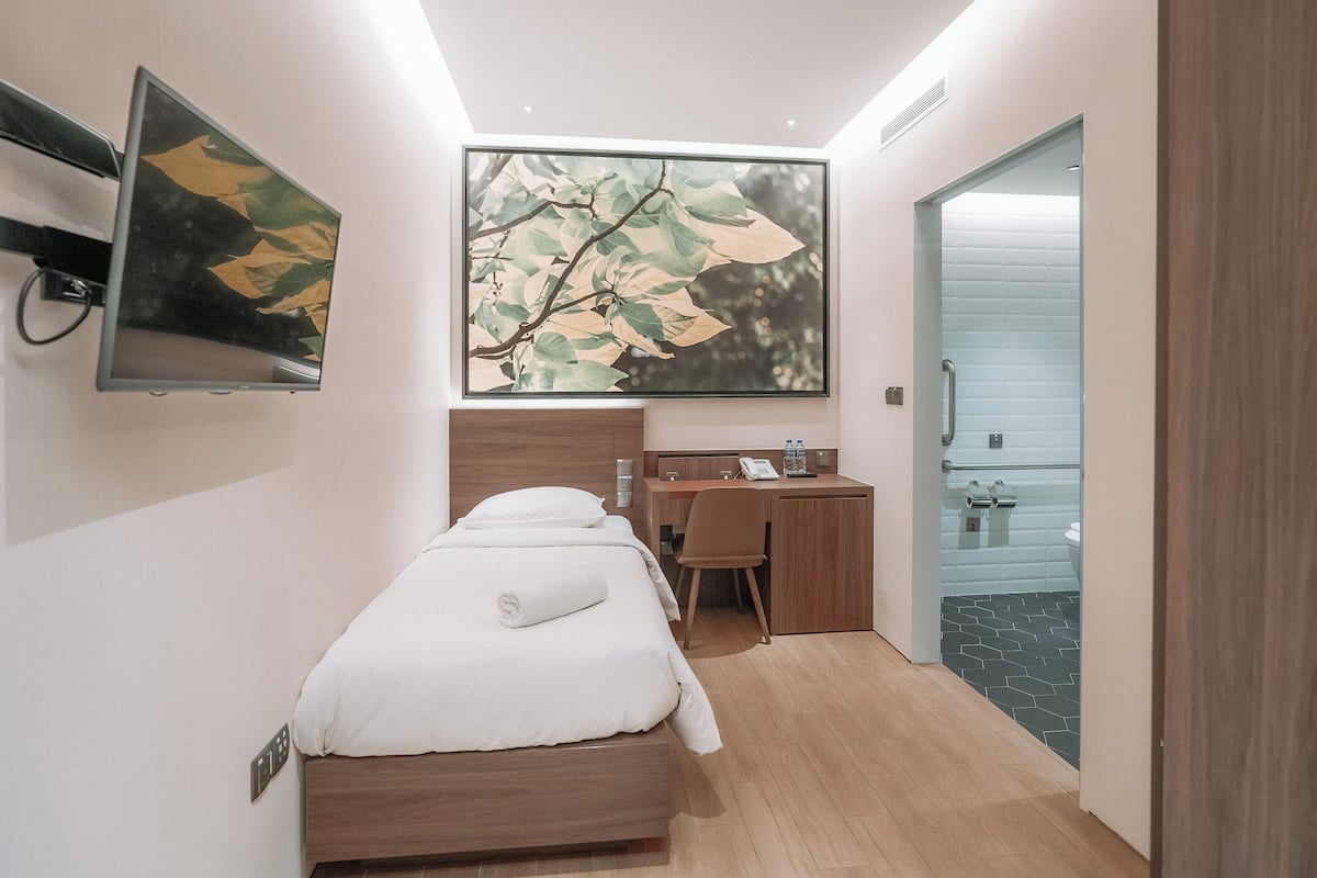 Quarto de hotel moderno com cama, escrivaninha e cadeira, uma janela grande e um banheiro visível por uma porta aberta.