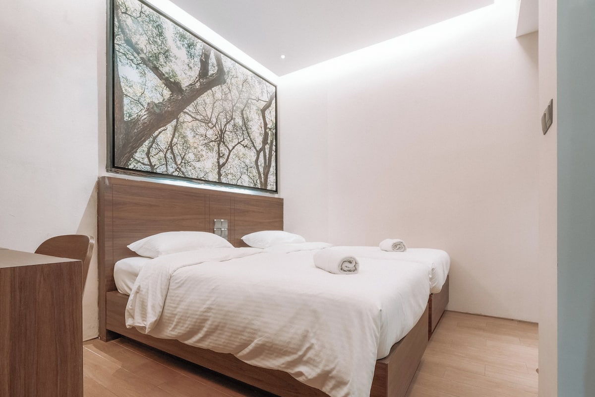 Quarto moderno com cama de madeira, roupa de cama branca e uma grande obra de arte com tema de árvore na parede.