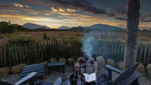 A rustic outdoor seating area with wooden benches and a stone fire pit, surrounded by a natural landscape and mountains in the background. - Romantic Rural Retreat - Rustic Cabin in the Mournes with Private Hot Tub (Annalong)