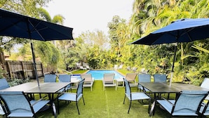 A poolside area with a rectangular pool, surrounded by chairs and tables under umbrellas, set against a backdrop of lush greenery. - Relaxing 2-Bedroom Apartment w / Pool Access (Fort Lauderdale)