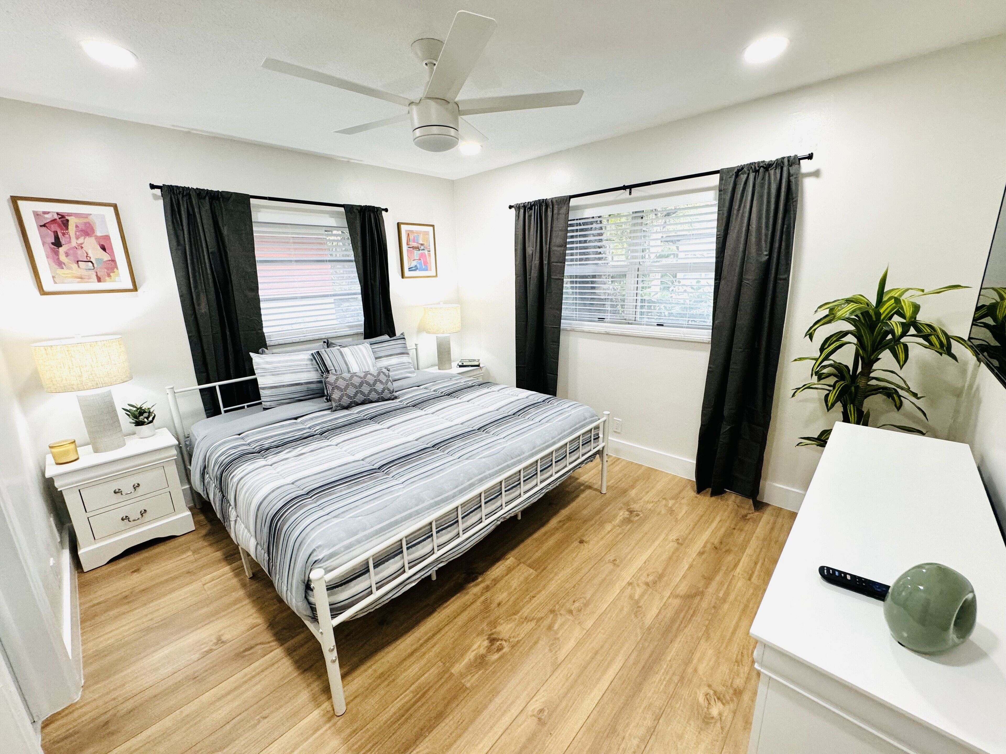A bedroom with a bed, bedside tables, a desk, a ceiling fan, and a potted plant.