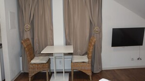 Une pièce avec une table blanche, deux chaises en osier, un téléviseur fixé au mur et des rideaux de couleur beige.