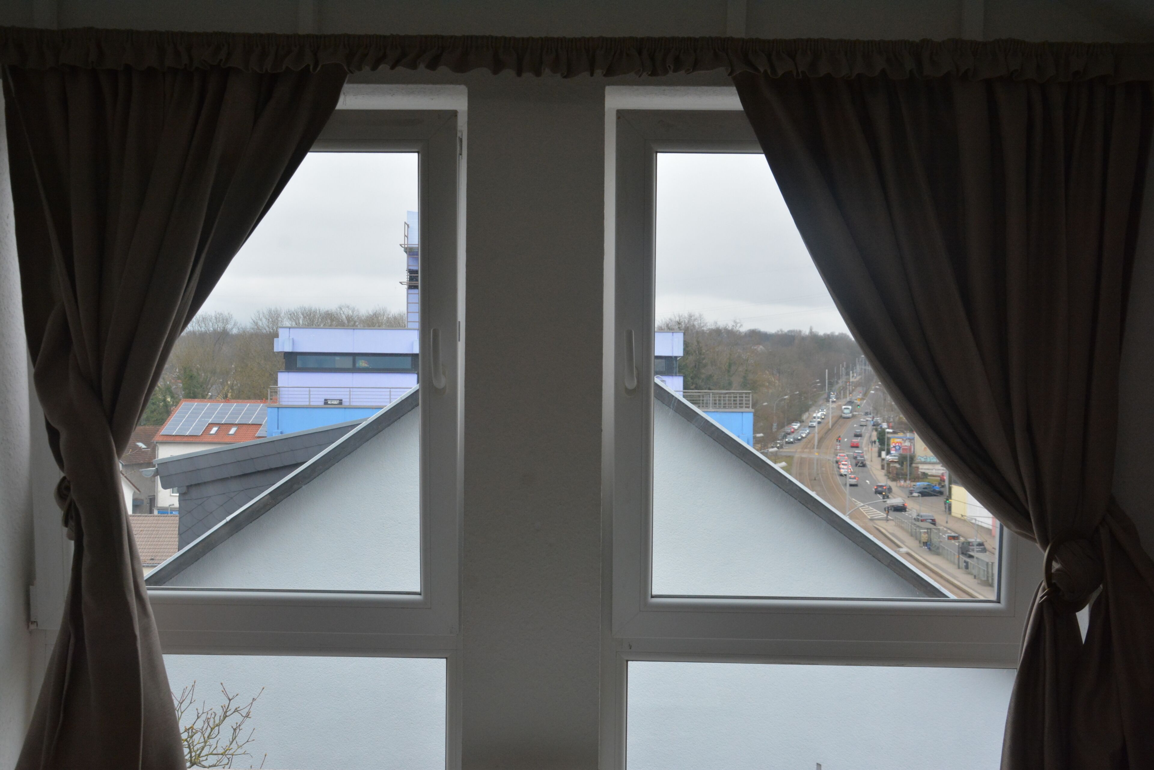 Ein Fenster mit Vorhängen, Blick auf eine Straße mit Fahrzeugen und Gebäude im Hintergrund.