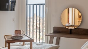 A hotel room with a bed, a small wooden table with a cup and saucer, a round mirror on a wooden shelf, and a view of a cityscape through a window with vertical blinds.