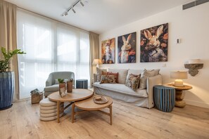 Een moderne woonkamer met een bank, een ronde houten salontafel en een groot raam met transparante gordijnen. Twee aan de muur hangende schilderijen van konijnen en een bijzettafeltje met een lamp.