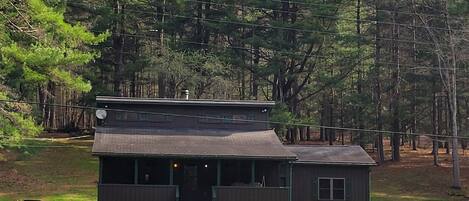 Uma cabana com varanda, mesa de piquenique e um pequeno edifício numa área arborizada.
