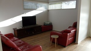 Una sala de estar con un televisor de pantalla plana sobre un mueble de madera, sofás de cuero rojo, una mesita redonda pequeña y una ventana con persianas.