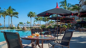 Un área de comedor junto a la piscina con mesas, sillas, sombrillas y palmeras.