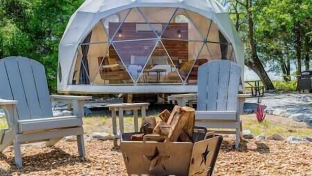 A geodesic dome structure with a wooden interior, surrounded by Adirondack chairs and a fire pit.