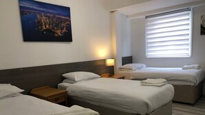 A hotel room with two beds, a nightstand, a lamp, and a window with blinds. - The Ullswater House  (Bournemouth)