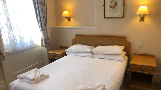 A neatly made bed with white linens and towels, two bedside tables with lamps, a framed botanical print on the wall, and a window with curtains. - The Ullswater House  (Bournemouth)