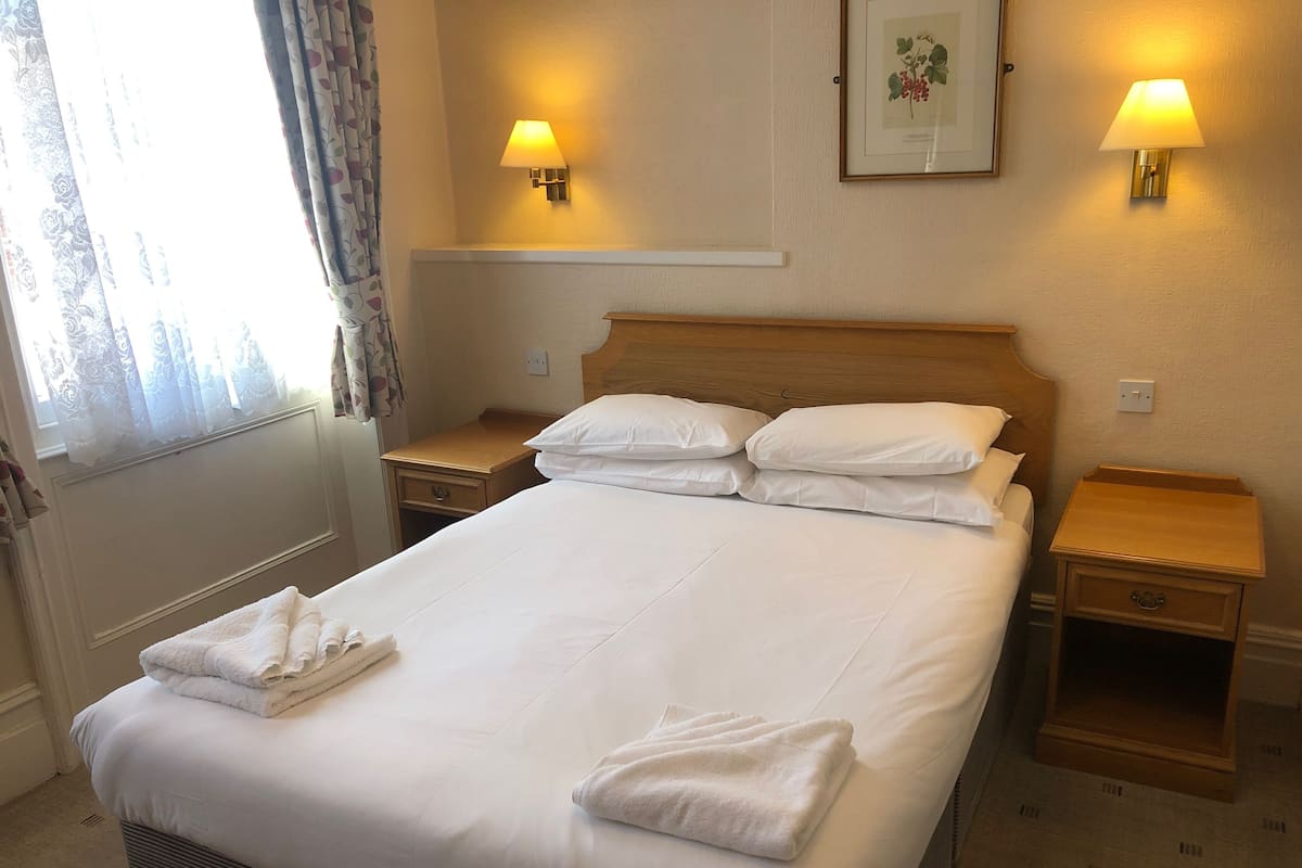 A neatly made bed with white linens and towels, two bedside tables with lamps, a framed botanical print on the wall, and a window with curtains.