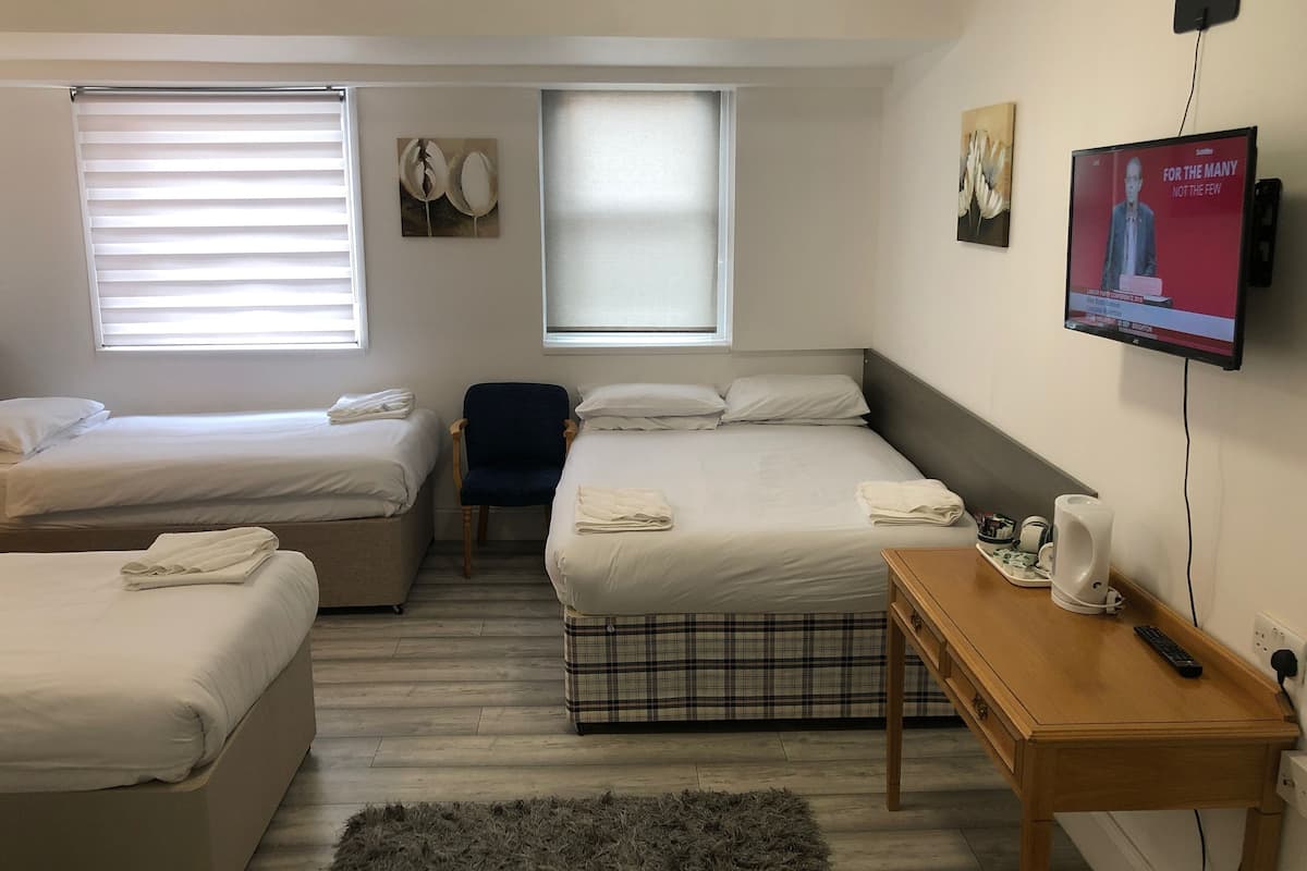 A hotel room with two beds, a chair, a wooden table with a kettle, a remote control, and a television displaying a news channel.