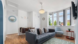 A modern living room with a grey sofa, a dining table with chairs, a round mirror, and a large window with a view of greenery. - Modern 4 bed 3 bath apartment, free parking (Coventry)