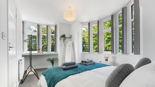 A bedroom with a large bed, a floor lamp, a mirror, and a view of greenery through large windows. - Modern 4 bed 3 bath apartment, free parking (Coventry)