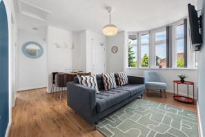Un salon moderne avec un canapé gris, une table à manger avec des chaises, une grande fenêtre avec des rideaux et une petite table d’appoint sur laquelle est posée une plante.
