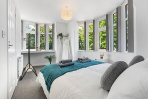 Et soveværelse med en stor seng, en gulvlampe, et spejl og udsigt til grønt over store vinduer.